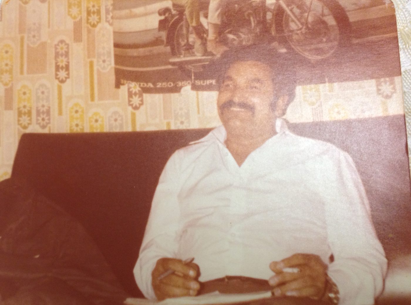 Mukhtar Ahmed smiling on a sofa in a white shirt, holding a pen and notebook